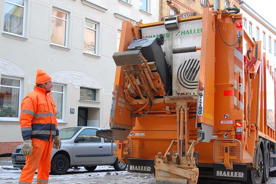 Ein Mann in orangefarbener Dienstkleidung steht neben einem Müllauto, an dem gerade eine Tonne geleert wird.