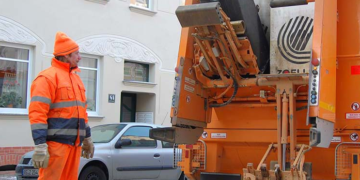 Ein Mann in orangefarbener Dienstkleidung steht neben einem Müllauto, in dem eine Mülltonne geleert wird.
