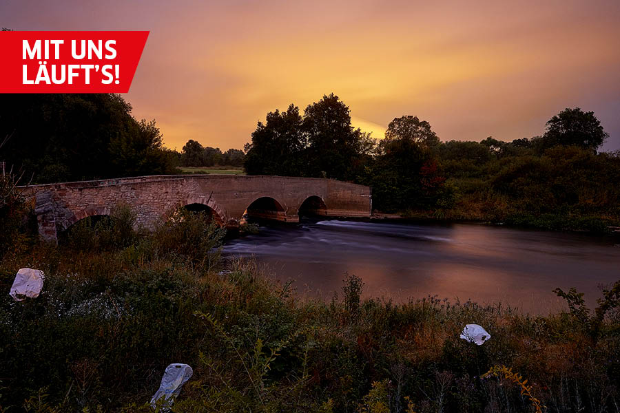 Zur goldenen Stunde wird die Schafbrücke in der Elsteraue im Süden der Stadt Halle (Saale) gezeigt. Es liegen Plastikbeutel auf den Grünflächen herum.