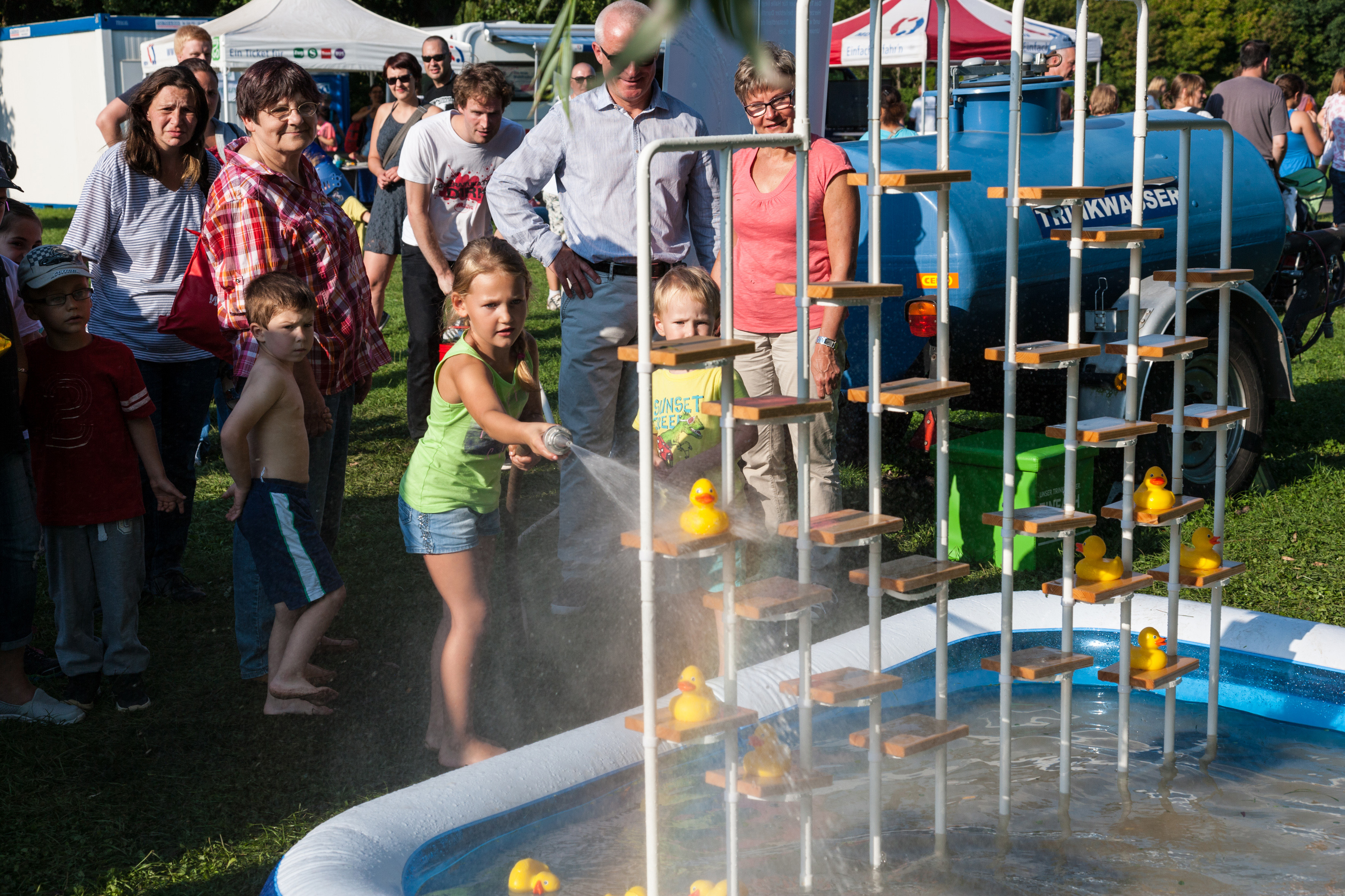 Kinder und Erwachsene stehen um einen blauen Pool. Ein Kind hält eine Wasserspritze in der Hand und versucht Enten von einem Regal zu schießen.