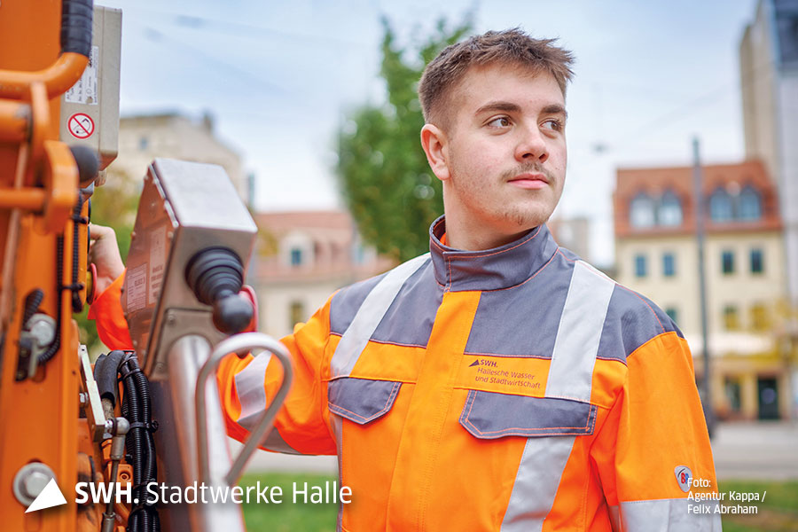 Ein junger Mann mit dunkelblonden Haaren schaut aus dem Bild heraus. Er trägt orangefarbene Arbeitskleidung der Halleschen Wasser und Stadtwirtschaft GmbH. Mit seiner rechten Hand bedient er ein Fahrzeug.
