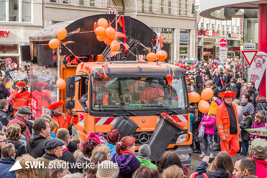 Ein orangefarbenes, großes Müllfahrzeug fährt mit Ballons und Girlanden behangen durch eine Menschenmenge.