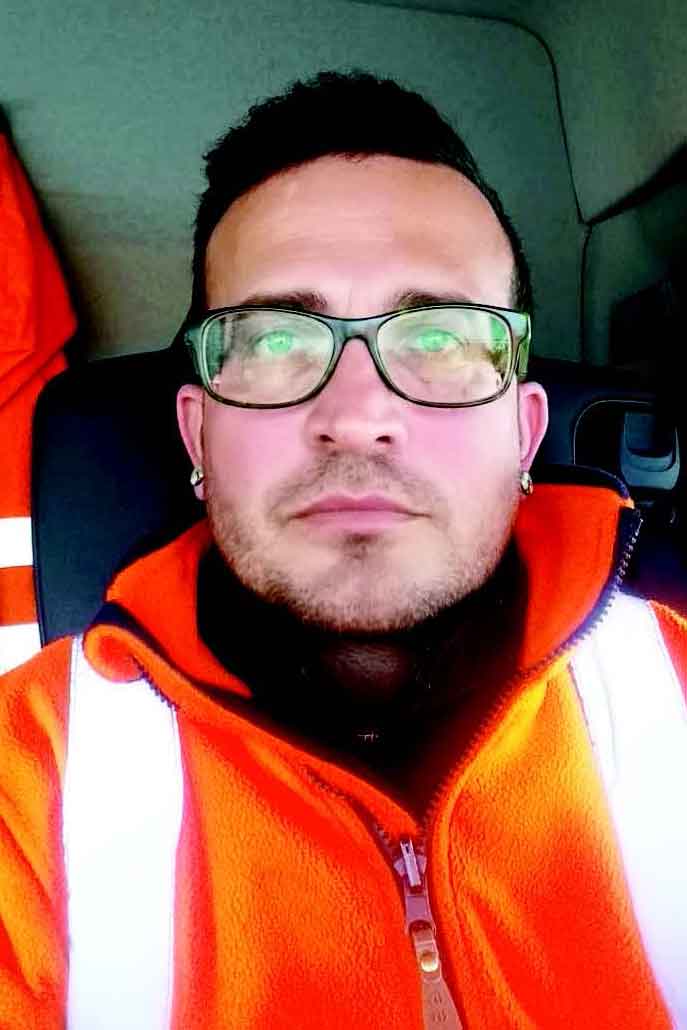 Das Foto zeigt das Gesicht eines Mannes, der in einem Fahrzeug sitzt und direkt in die Kamera schaut. Er hat dunkles Haar und einen Bart, trägt eine Brille und ein orangefarbenes Oberteil.