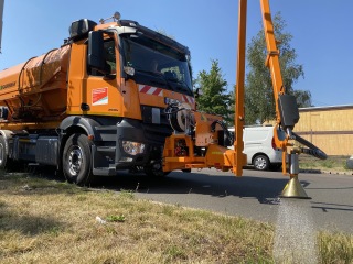 Das Foto zeigt ein großes orangefarbenes Fahrzeug, an dessen Front eine Art Arm befestigt ist, mit dem Bäume bewässert werden können.