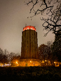 In der Bildmitte befindet sich der Wasserturm Süd, ein zehneckiger Turm mit braun-roten Klinkersteinen. Der Fuß, sowie die oberste Etage des Turms werden in warmen orangefarbenen Tönen beleuchtet. Um den Turm herum ist der Himmel grau und die Bäume neben dem Turm wirken im Dämmerlicht schwarz.